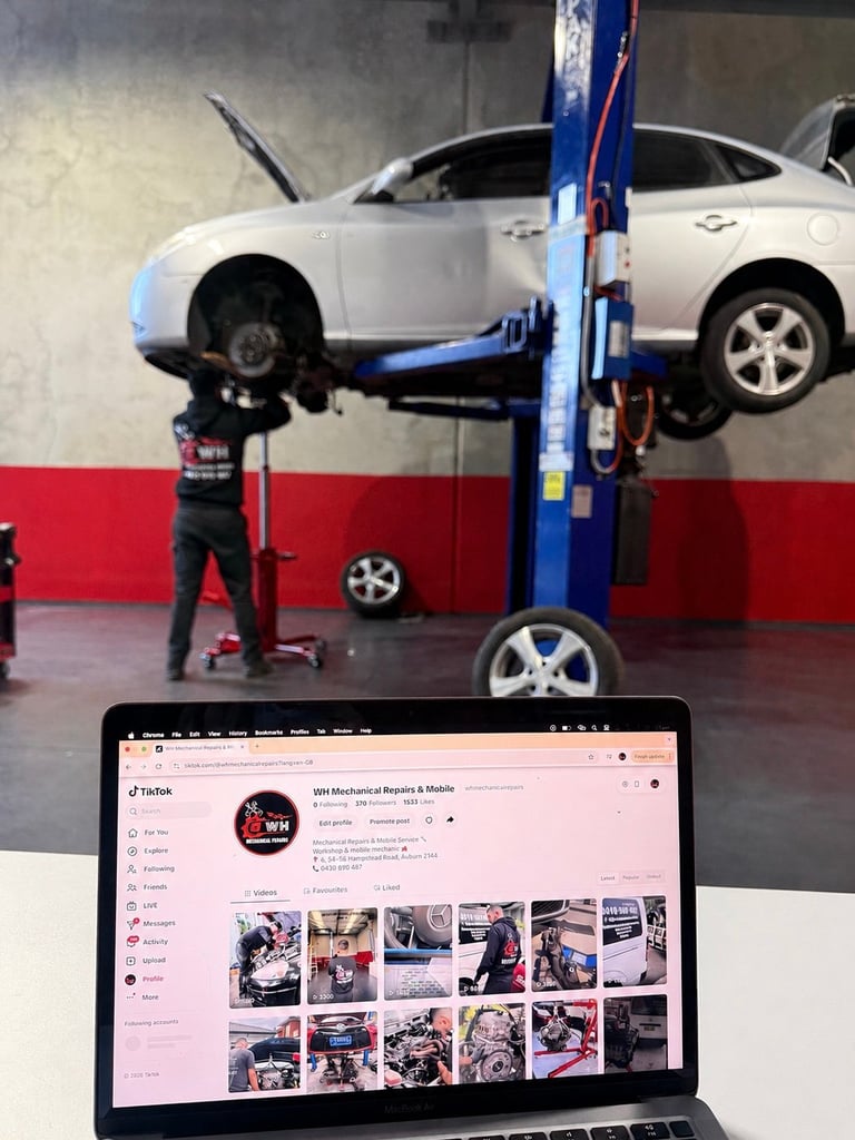 Auto repair shop with white car on blue hydraulic lift, mechanic working underneath, laptop displaying social media in foreground