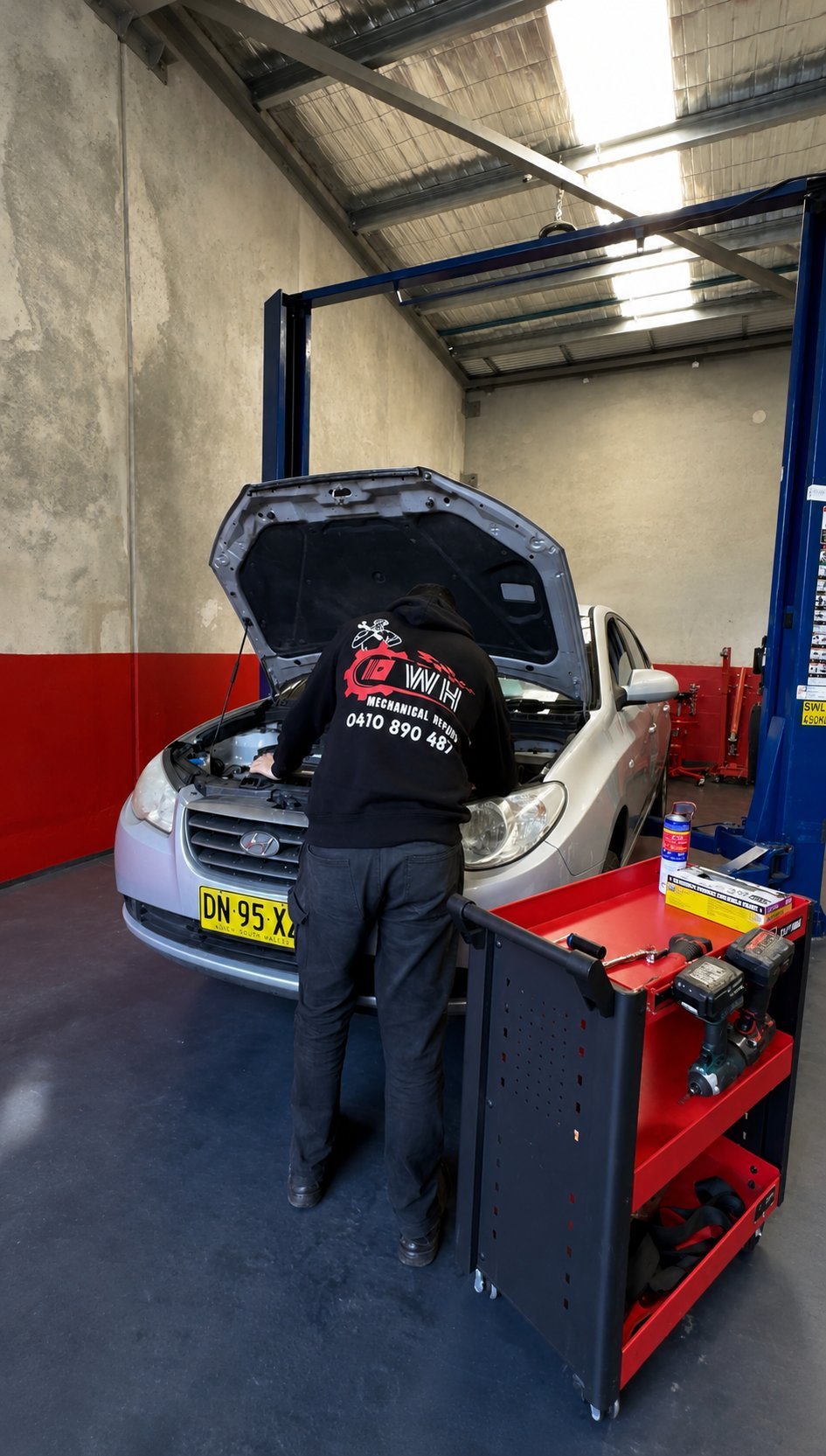 Mechanic inspecting engine of silver car in workshop with red tool cabinet and metal lift structure